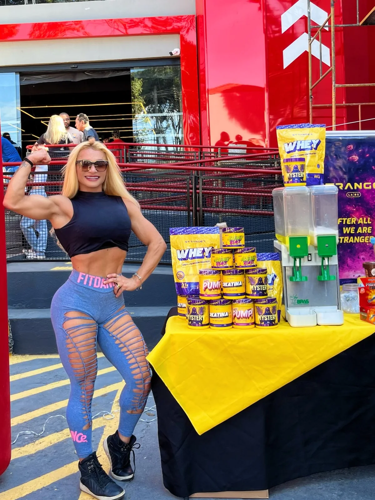 Evelyn Soares flexionando o braço ao lado de uma mesa de suplementos em um evento